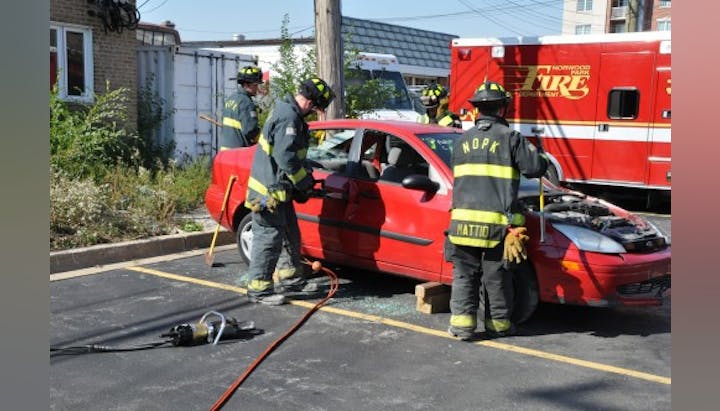 Firefighters in gear practice removing a car door next to a red vehicle in a parking lot, with a fire truck in the background.