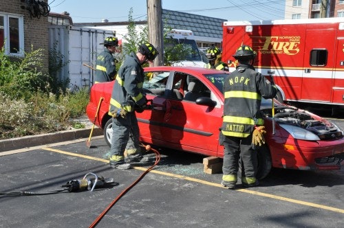 Firefighters in gear practice removing a car door next to a red vehicle in a parking lot, with a fire truck in the background.
