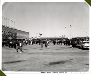 A vintage photo of people walking in a parking lot, with cars and a building in the background.