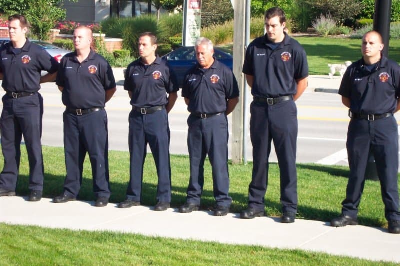 Seven uniformed individuals stand in a line on a sidewalk, likely firefighters, outdoors in a grassy area.