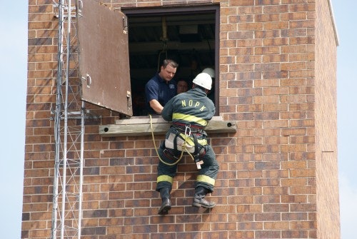 A firefighter in gear is climbing into a building through a window with assistance from another person inside.