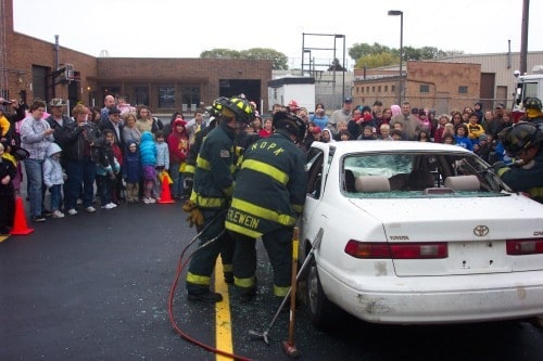 Firefighters demonstrating vehicle extraction techniques to a crowd using a white car.