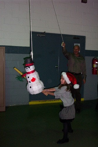 A child in a Santa hat hits a snowman piñata with a bat while an adult holds the rope indoors.