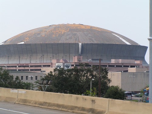 A large, weathered domed stadium with visible wear on the roof, surrounded by trees and nearby buildings.