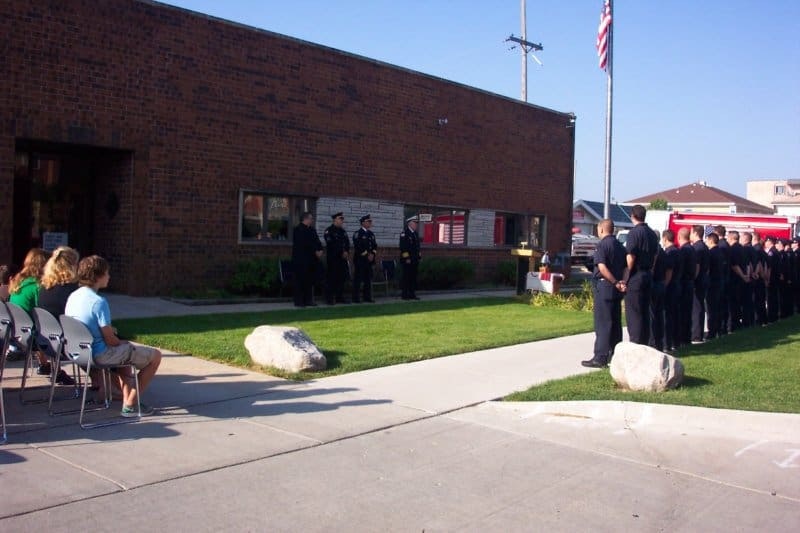 A group of uniformed individuals stands in formation outside a brick building, with spectators seated nearby. An American flag is visible.