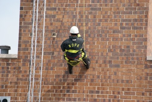 A firefighter is rappelling down a brick wall in gear, wearing a helmet and harness.