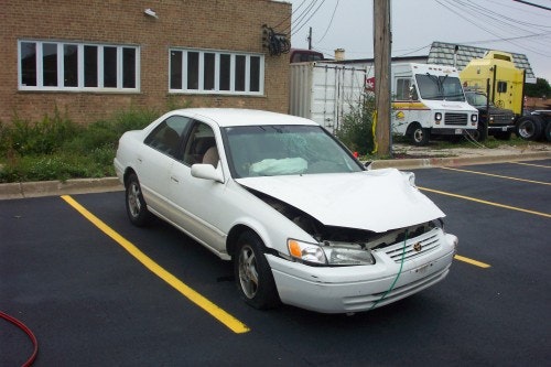 A white car with front-end damage is parked in a lot.