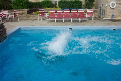 A splash in a swimming pool with red lounge chairs and greenery in the background.