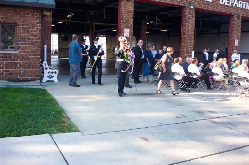 A gathering outside a brick fire department with people seated and standing, some in uniform holding a floral wreath.