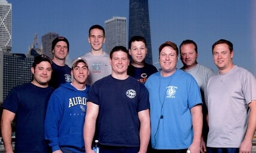 A group of nine men posing in front of a cityscape background wearing casual and sports-themed clothing.