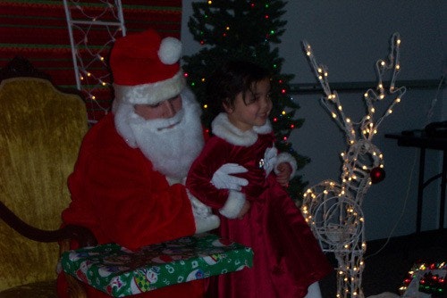 Santa with a child on his lap, next to a lit reindeer and Christmas tree.