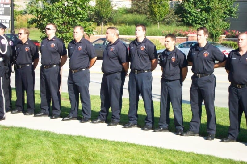 A group of uniformed firefighters standing in a line outdoors on a sunny day.