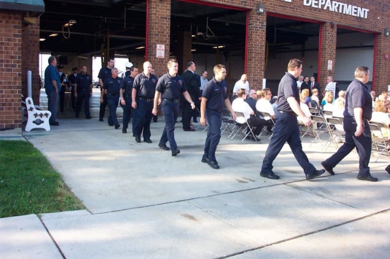A group of people in uniform walking past seated audience outside a fire department.