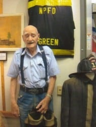 An elderly man stands smiling near a firefighter's uniform and helmet. There's a sign labeled "NPFD GREEN" on the wall.