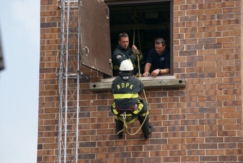 A firefighter is rappelling down a building while two people assist from a window above.