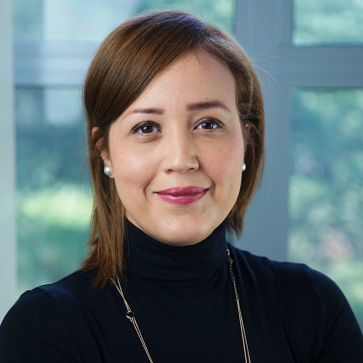 A portrait of a woman with brown hair and earrings, wearing a black turtleneck, smiling in a bright indoor setting.