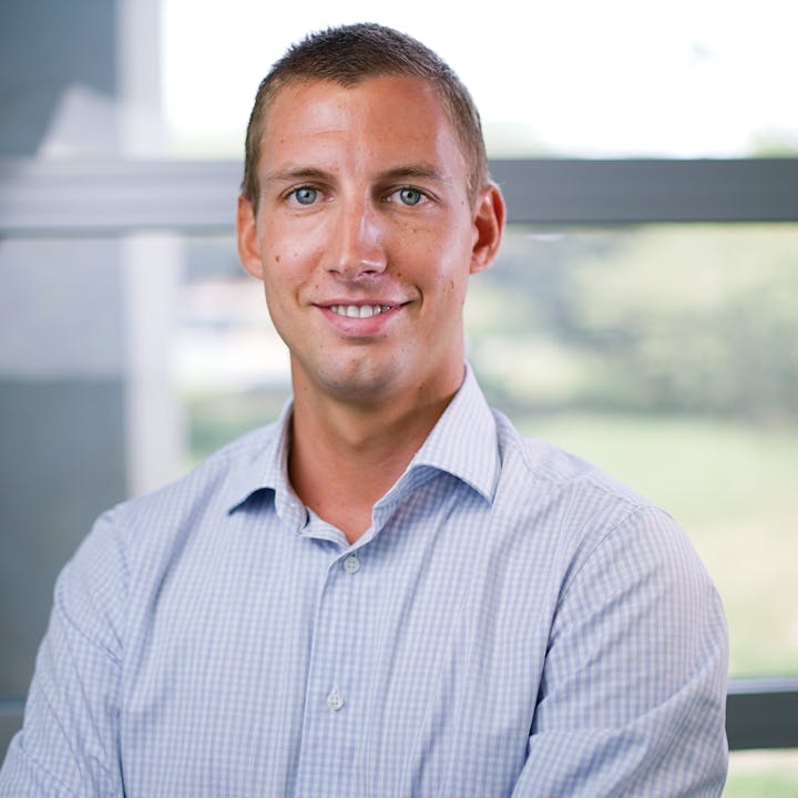 A man in a light blue checkered shirt smiles confidently, standing in front of a window with greenery outside.