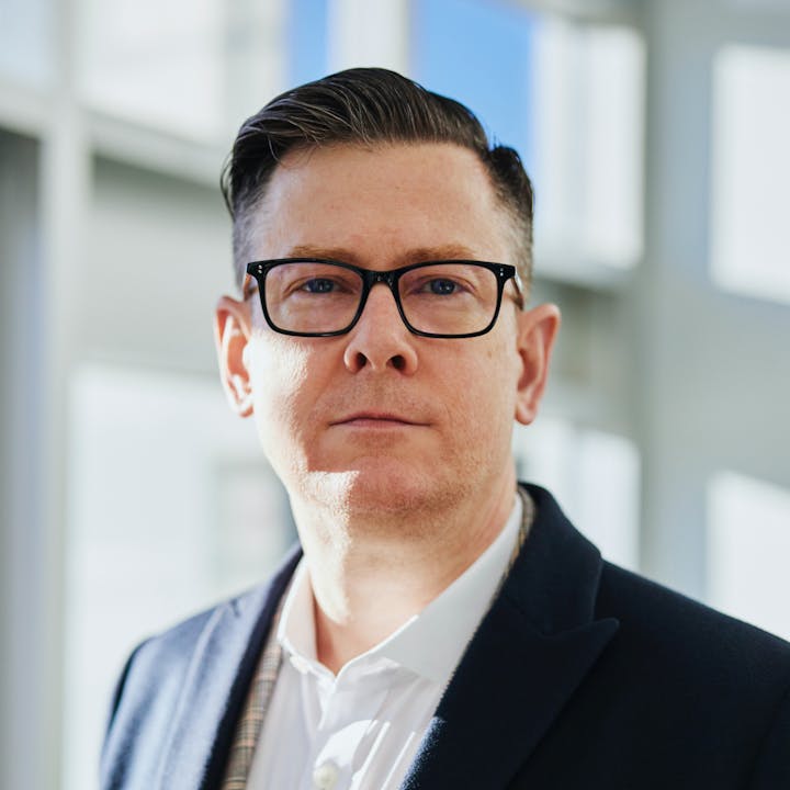 A person with glasses looks confidently at the camera, wearing a blazer and a collared shirt, in a well-lit indoor setting.
