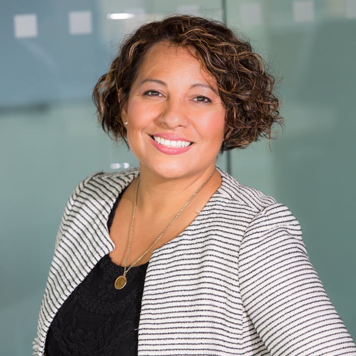 A smiling woman with curly hair, wearing a striped blazer over a black top, poses confidently in a modern office setting.