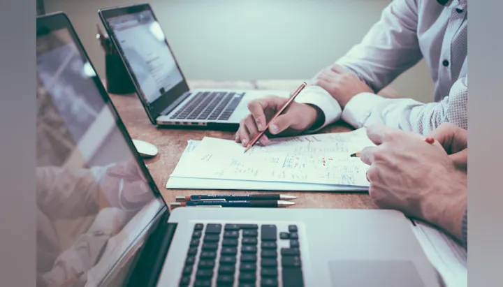 Two people are collaborating over documents at a desk with laptops, pens, and a cup, discussing or planning something together.