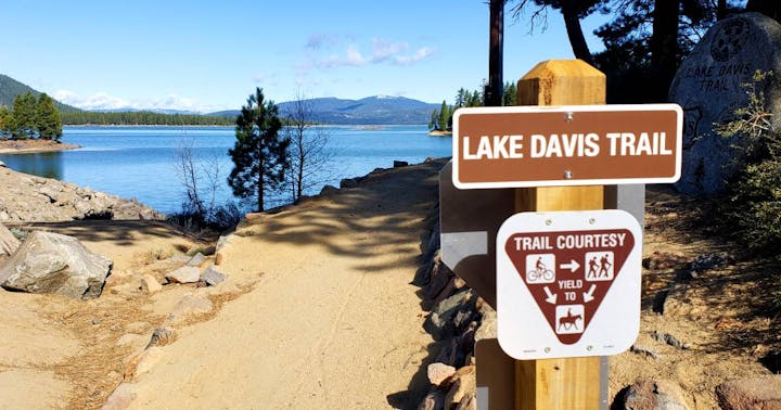 Lake Davis Trail sign near a lake with mountains in the background, advising trail courtesy.