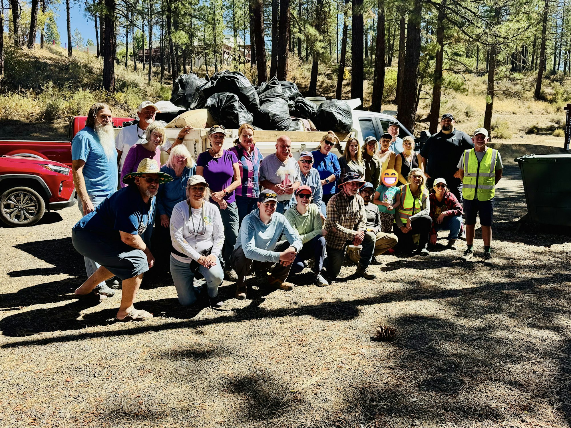 A group of people poses together outdoors, surrounded by trees, with a truck loaded with bags behind them.