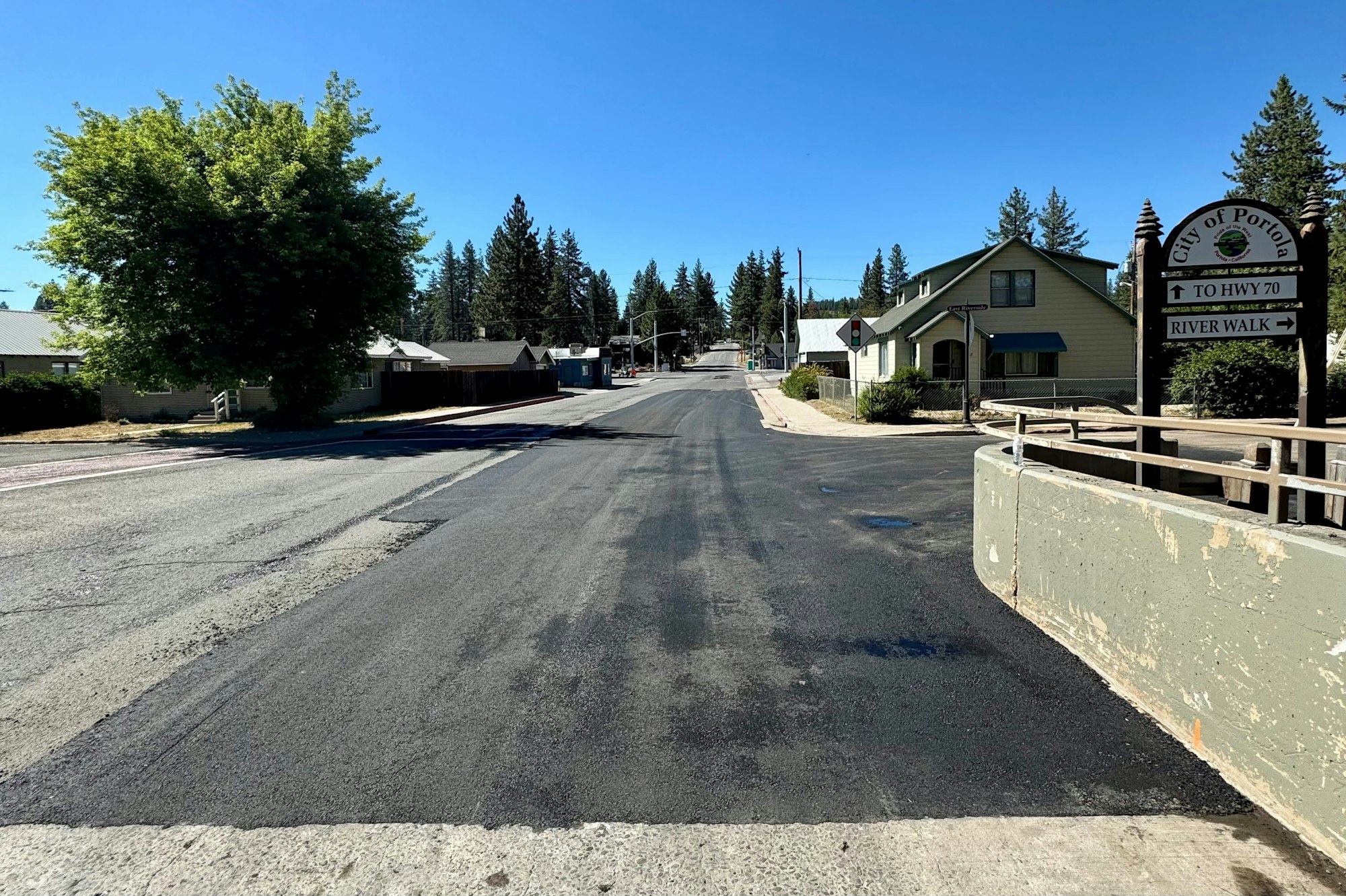 A road in a small town with houses, trees, and a sign pointing to "TO HWY 70" and "RIVER WALK" under a clear blue sky.