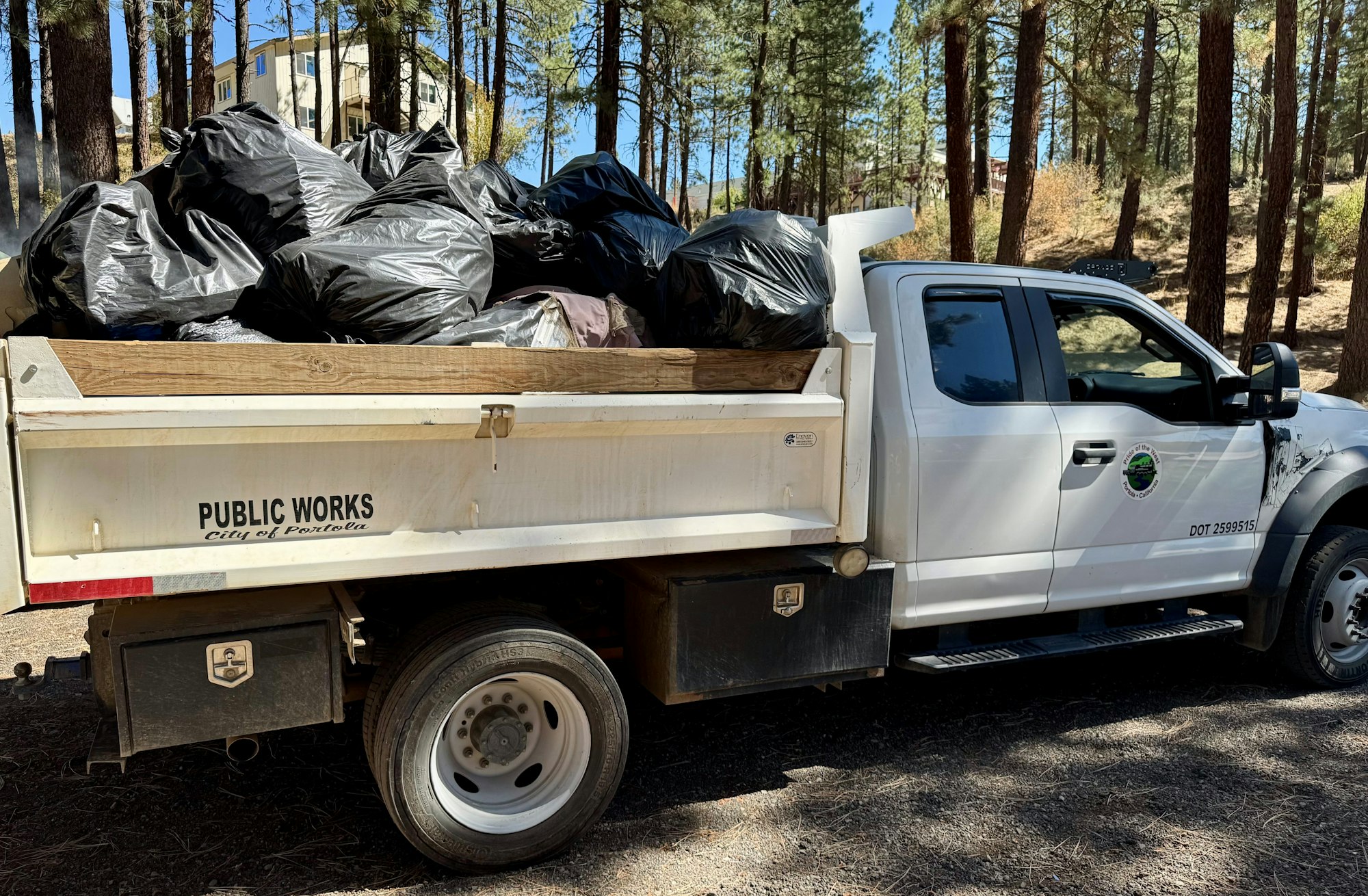 A truck from the Public Works department is loaded with black garbage bags, parked in a forested area.