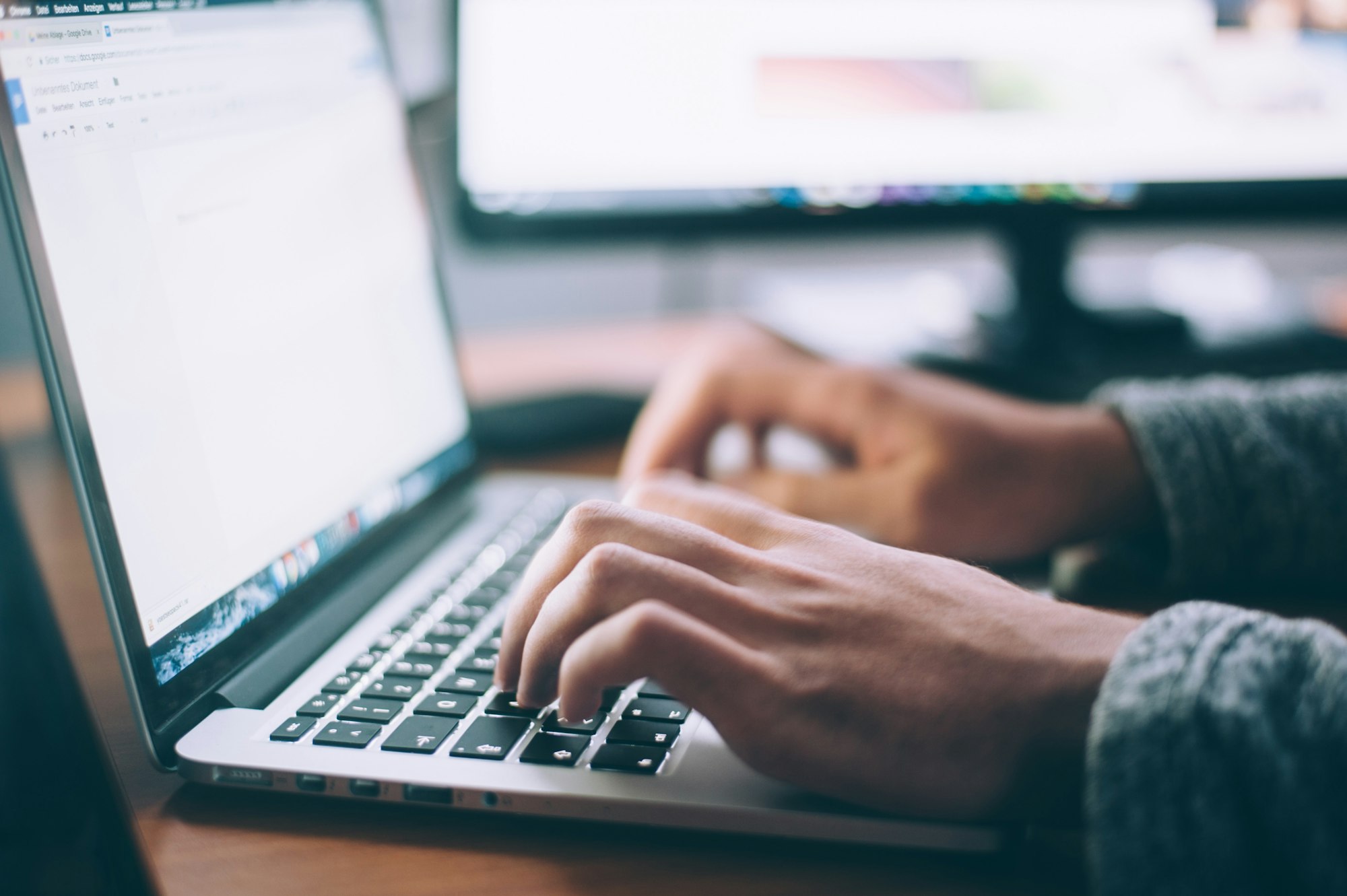 Close-up of hands typing on a laptop keyboard with a blurred screen and second monitor in the background.