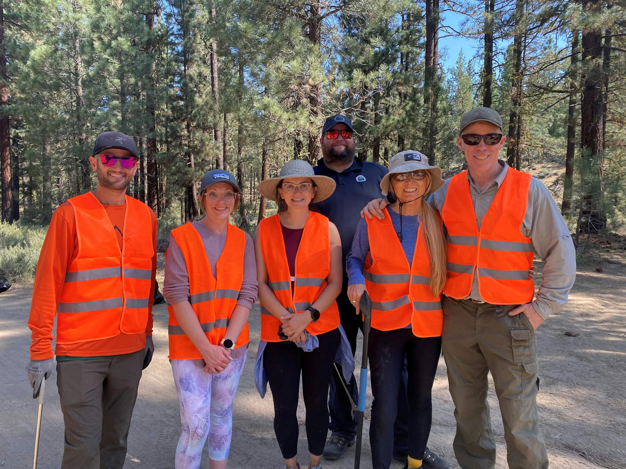 Six people in bright orange vests pose in a forest setting, suggesting an outdoor or environmental activity.