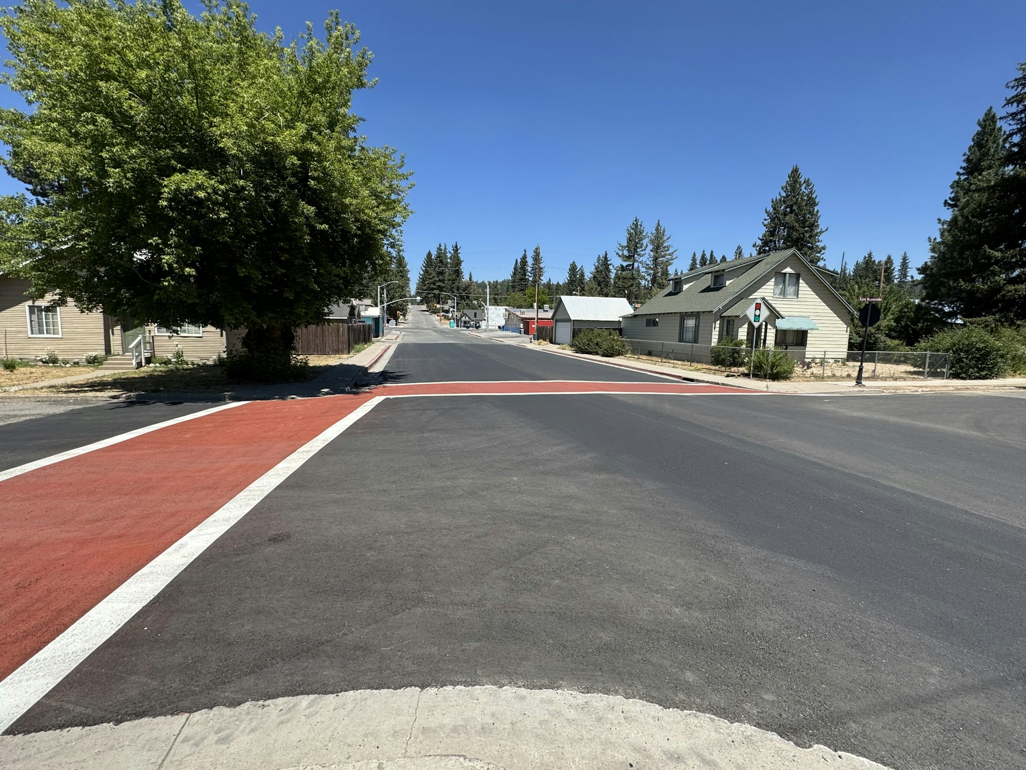 Road intersection with a red crosswalk, houses, trees, and a clear blue sky.