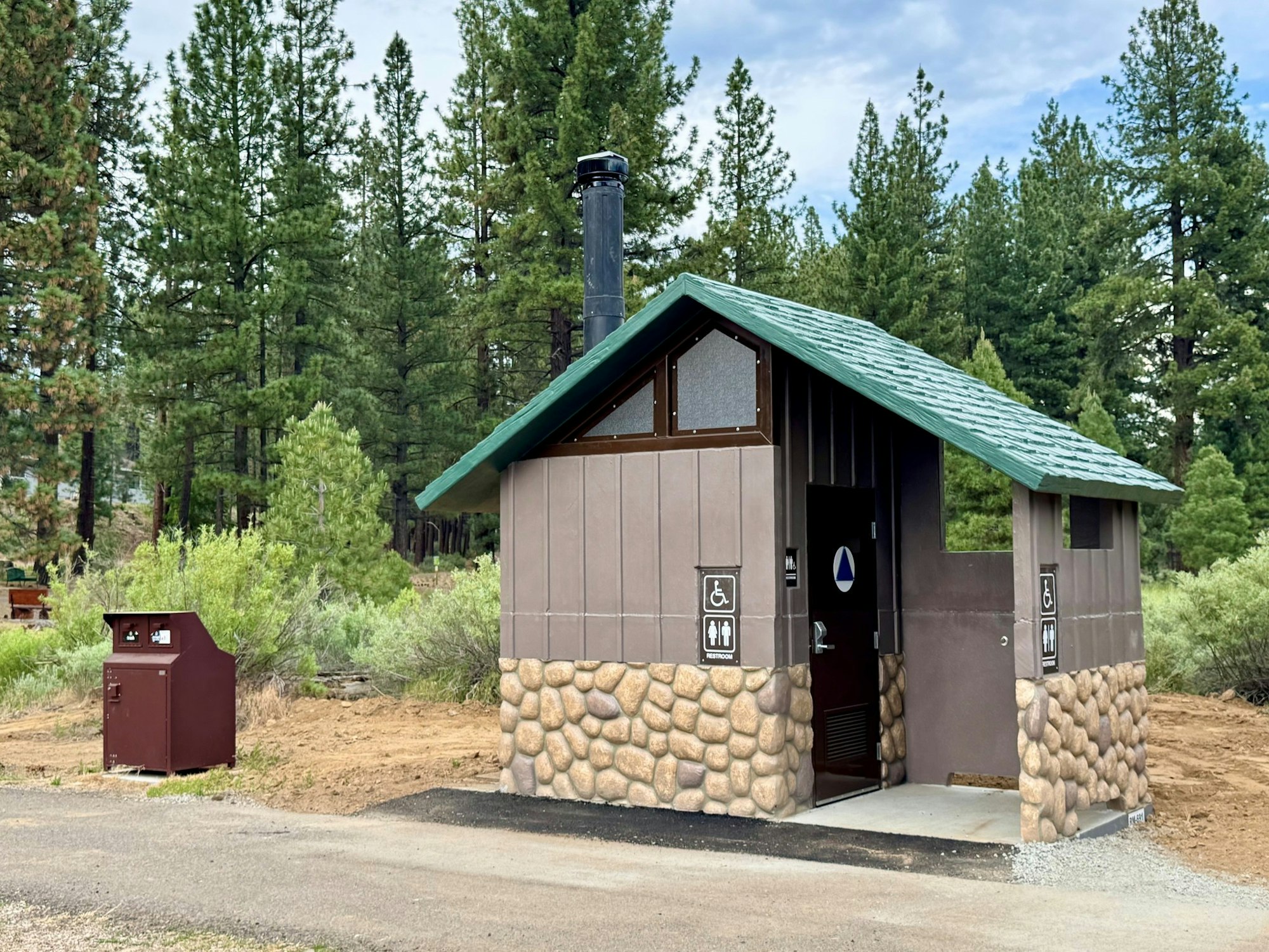 Small restroom building with disability access sign and trash bin, set in a forested area.