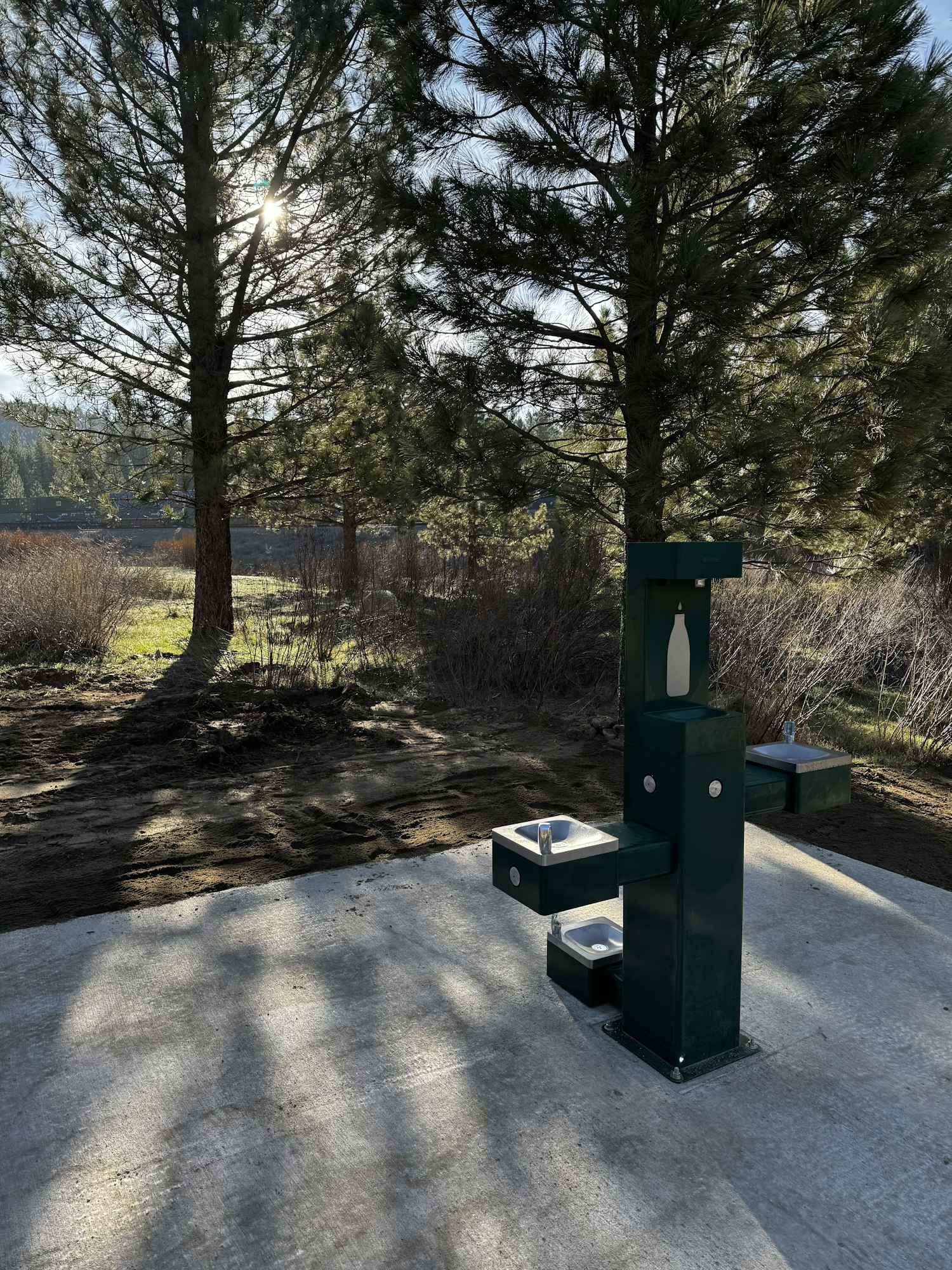 The image shows a green water fountain with two drinking stations, surrounded by trees and a sunny outdoor setting.