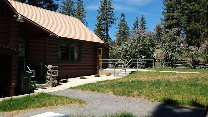 A log cabin with a green lawn, trees, clear skies, and a walking path.