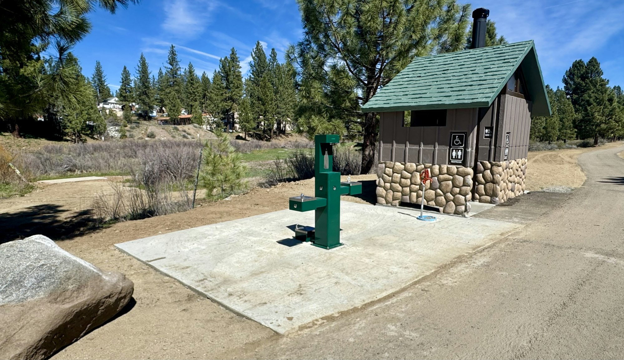 The image shows a restroom building with a stone base and a green water fountain nearby, surrounded by trees and a dirt path.