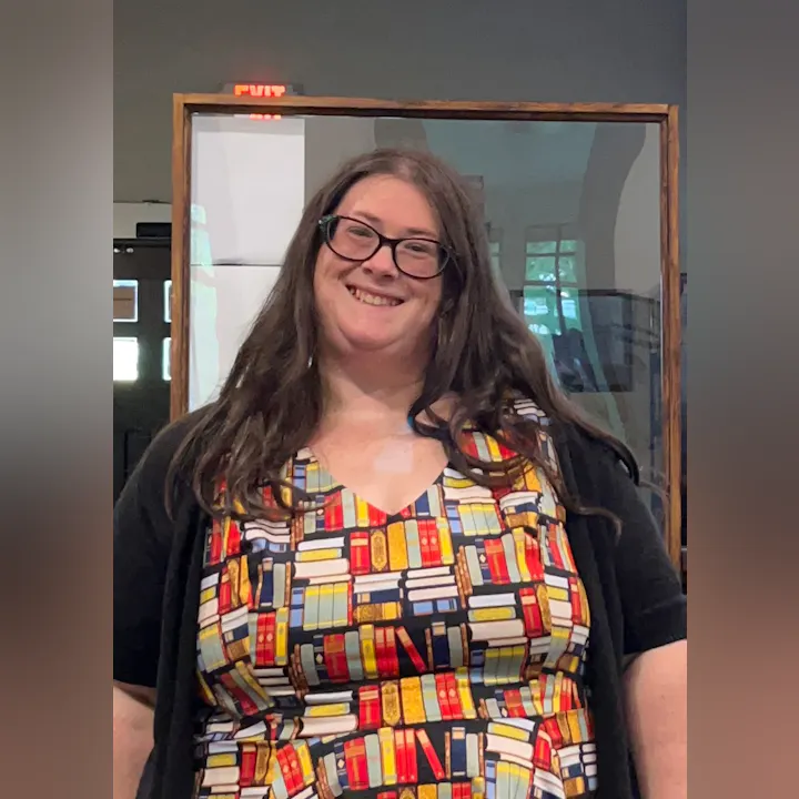 A smiling person with glasses wearing a colorful top with bookshelf pattern, standing indoors near an EXIT sign.