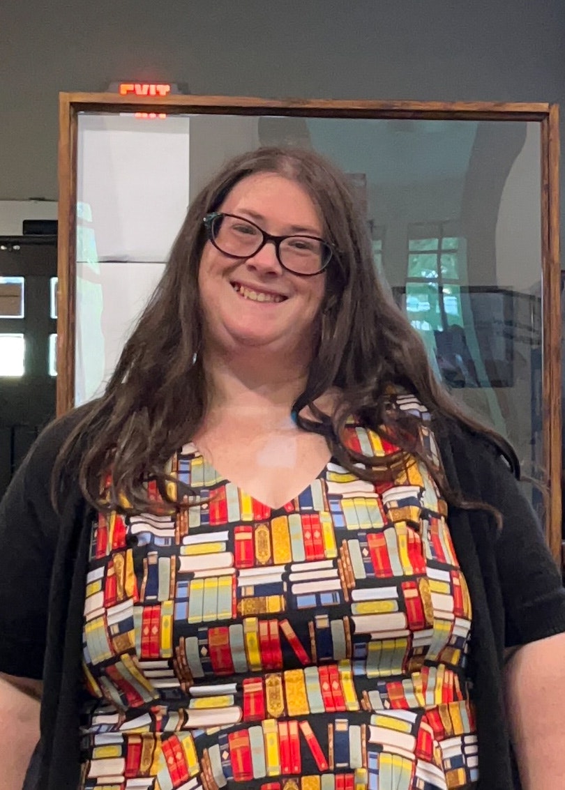 A smiling person with glasses wearing a colorful top with bookshelf pattern, standing indoors near an EXIT sign.