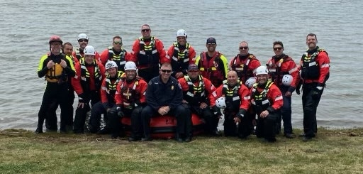 A group of 20 rescue workers in red and black gear pose by a body of water, showcasing teamwork and readiness for rescue operations.