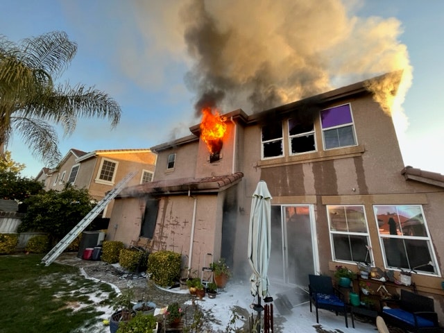 A house is on fire, with flames and smoke billowing from the upper windows, and firefighters' equipment visible nearby.
