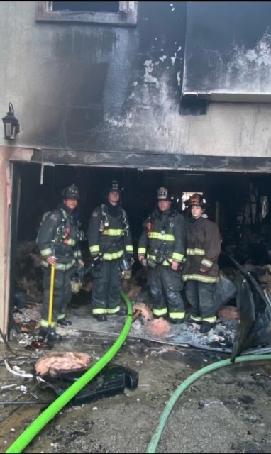 Four firefighters stand in front of a burned building, with debris around them and a hose visible on the ground.