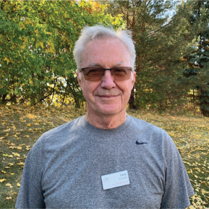 A smiling man in a grey shirt with a name tag, standing outside with trees and fallen leaves in the background.