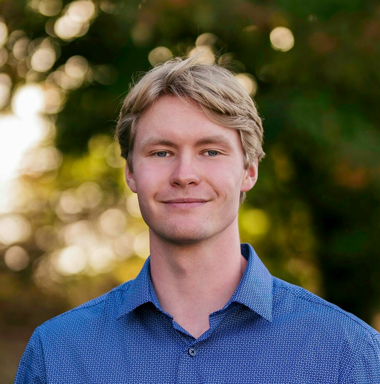 A person wearing a blue shirt stands outdoors with a blurred green background.