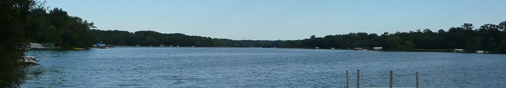 A panoramic view of a calm lake with surrounding trees and docks, under a clear sky.
