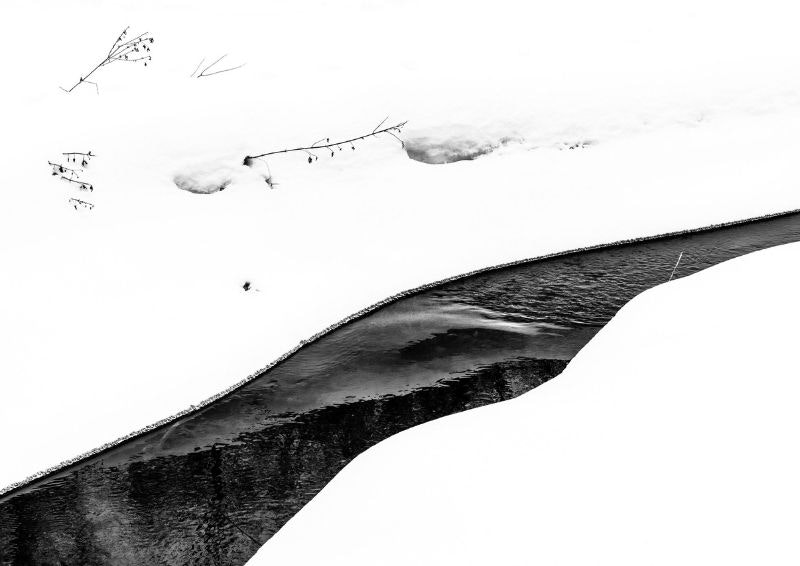 Snow-covered landscape with a curving stream and bare branches poking through the snow.