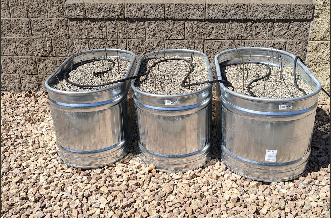 Three metal raised garden beds with gravel and drip irrigation systems, against a brick wall on a pebble-covered ground.