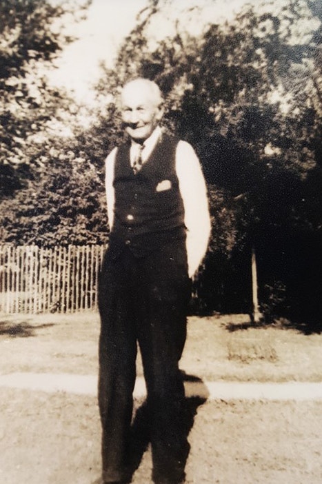 Elderly man in a vest and trousers, smiling, in a sunny garden with a fence and trees. Vintage black and white photo.