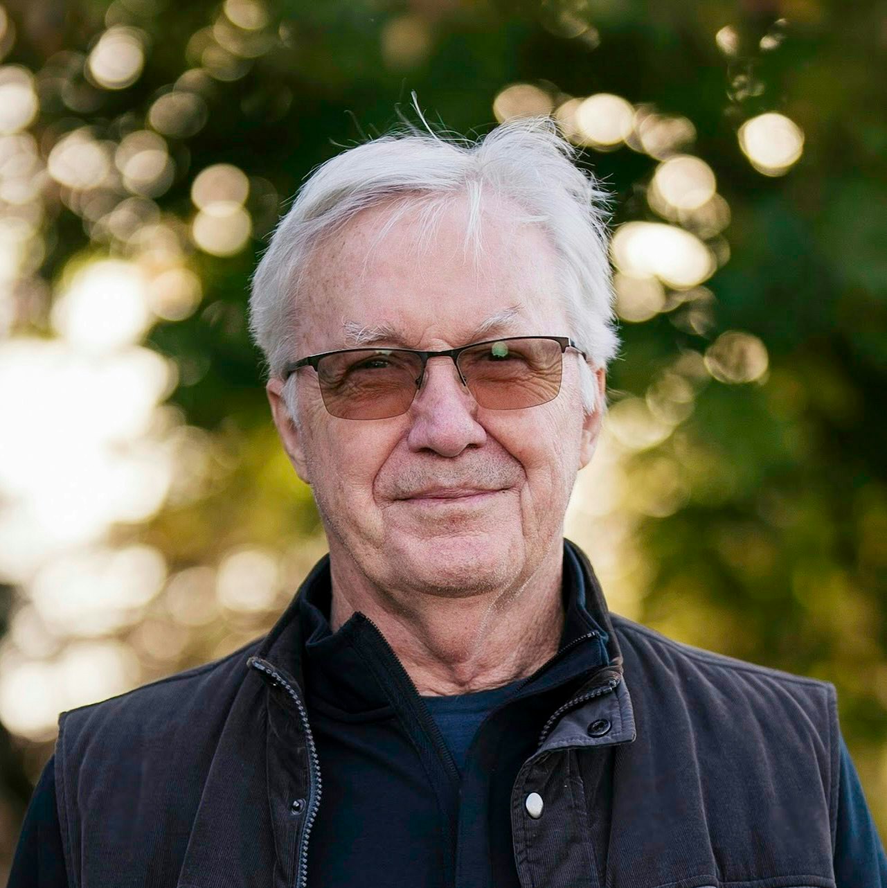 An older man with glasses and white hair, wearing a dark jacket, stands outdoors with a blurred leafy background.