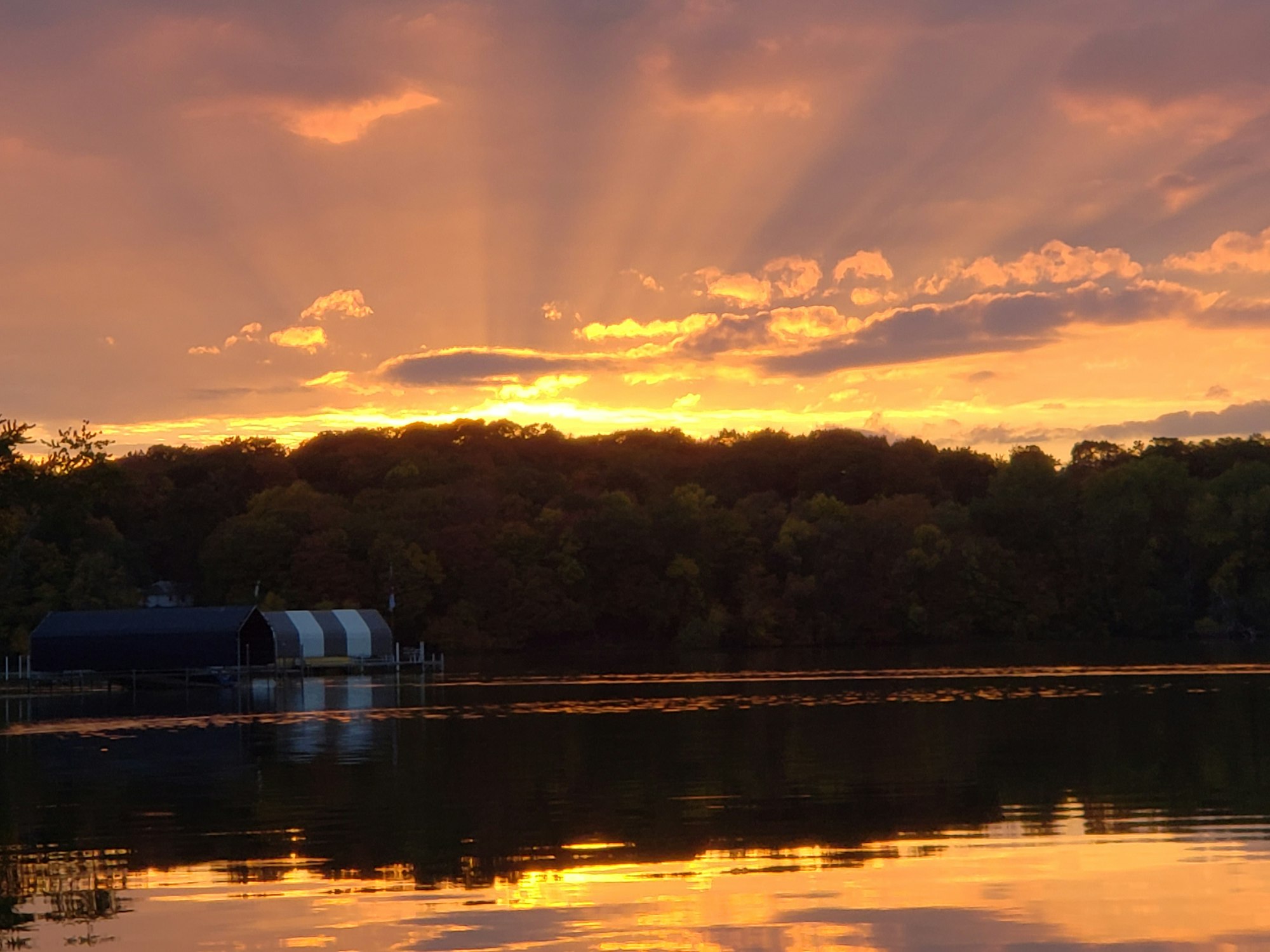 A vibrant sunset over a calm lake with trees and a boathouse. Sun rays pierce through clouds, casting reflections on the water.