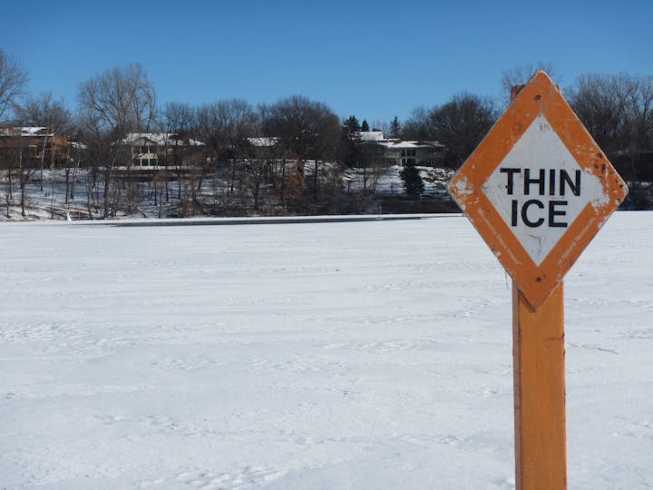 A "THIN ICE" warning sign beside a snowy, icy surface with trees and houses in the background.