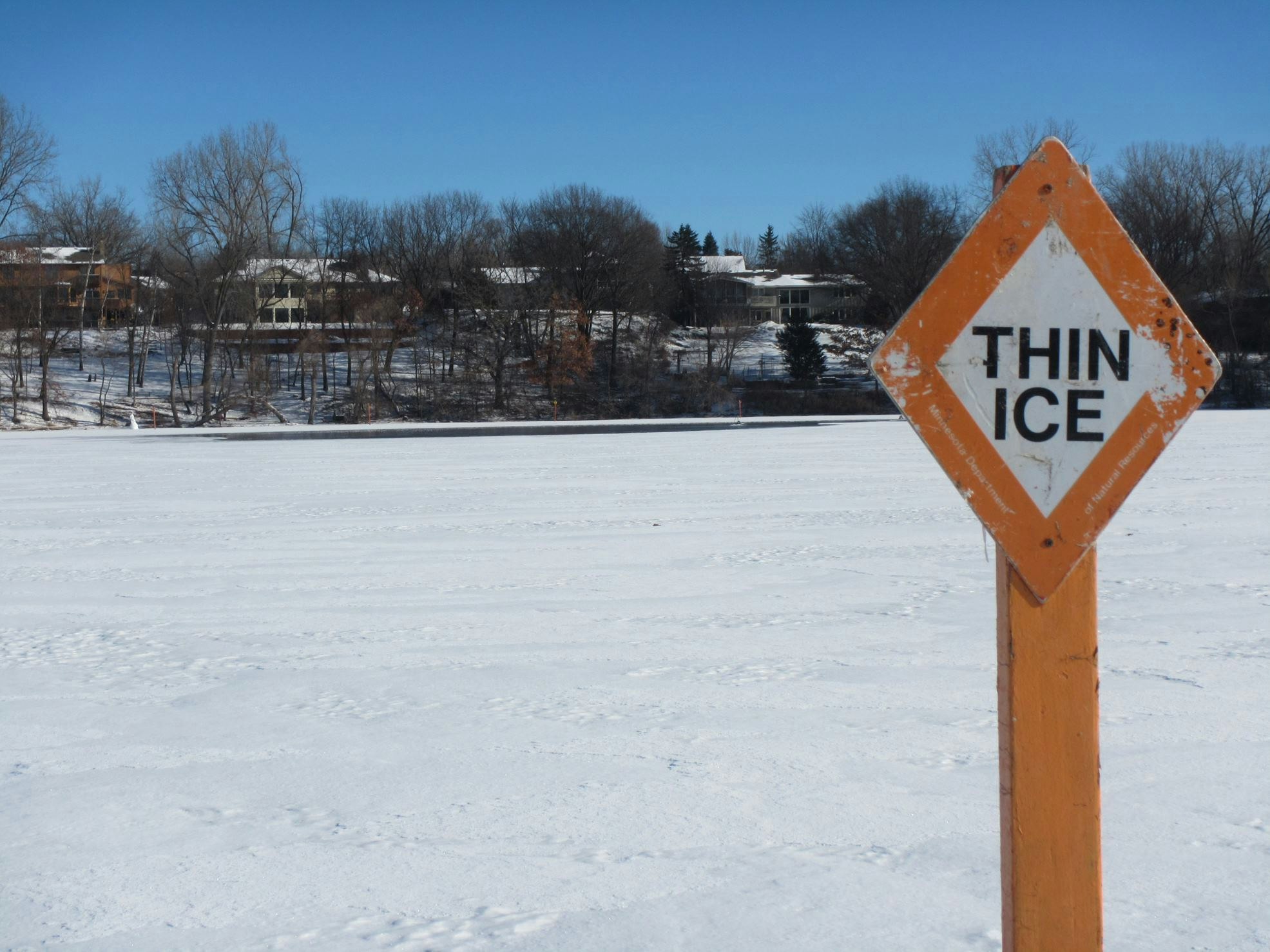 A "THIN ICE" warning sign beside a snowy, icy surface with trees and houses in the background.